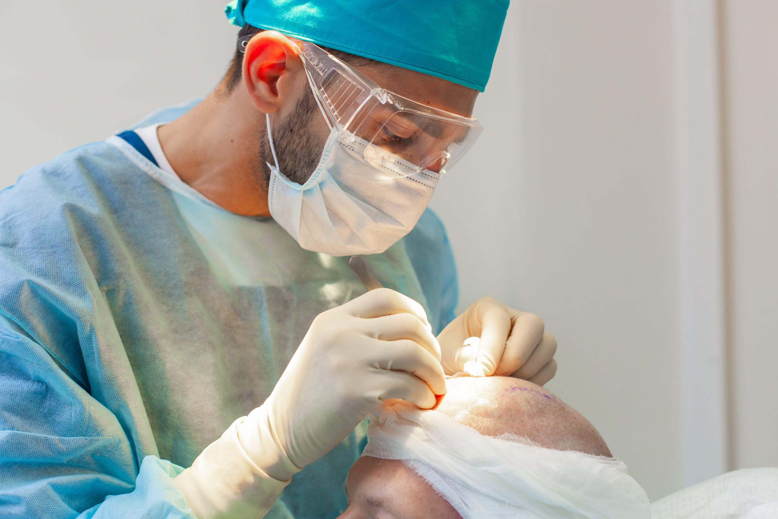 Doctor in scrubs and surgical mask performing a hair transplant procedure on a male patient.
